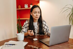 Woman in wireless headphones looks at laptop with doubt and poses in office